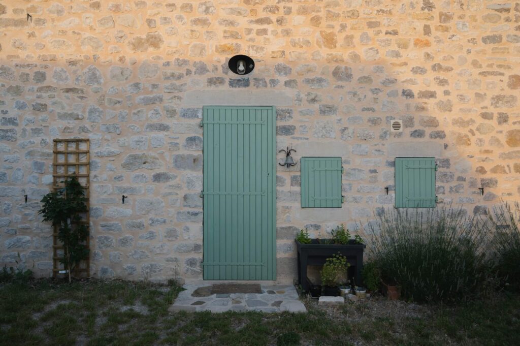 Rustic French-style stone cottage wall with a pastel sage green wooden door and shutters, embodying the quiet, grounded strength of those who embrace femininity. A small climbing rose trellis and lavender bushes line the path, symbolizing the gentle unfolding of the self.
